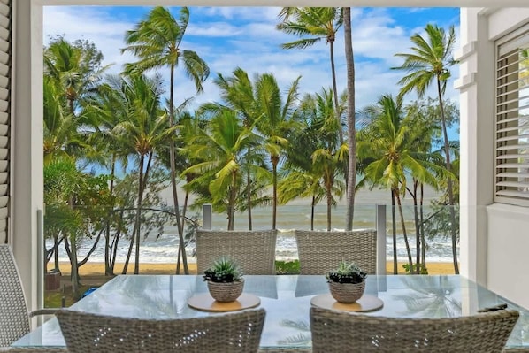 A balcony with wicker furniture, a glass table, and a view of the beach and palm trees.