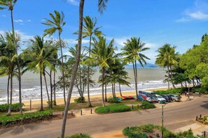 A beachside view with palm trees, a road, parked cars, and a bus.
