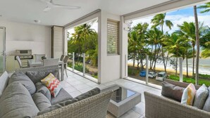 A modern living room with a sofa, chairs, a coffee table, and a view of the beach and palm trees. - Mistral on Island Views—Pool Paradise on the Beach (Palm Cove)