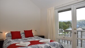 Ein Schlafzimmer mit einem Bett, zwei roten Kissen, einer weißen Decke und Blick auf einen Balkon mit Sitzgelegenheit.