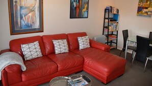 Ein rotes Ledersofa mit geometrischen Kissen, ein Glastisch mit Zeitschriften und ein Bücherregal mit Büchern und einem Laptop.