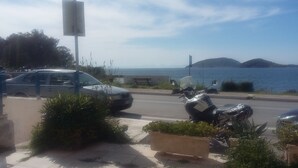 Une route côtière bordée de voitures garées et d’un scooter, avec un banc et une vue sur la mer et les collines.
