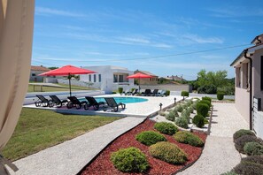 Zona de piscina con sillas de descanso y sombrillas rojas, rodeada de jardines cuidados y un cielo despejado.