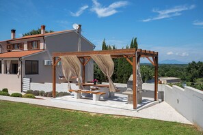 Een huis met een patio, voorzien van een houten pergola met een zonnedak, een bank en een tafel.