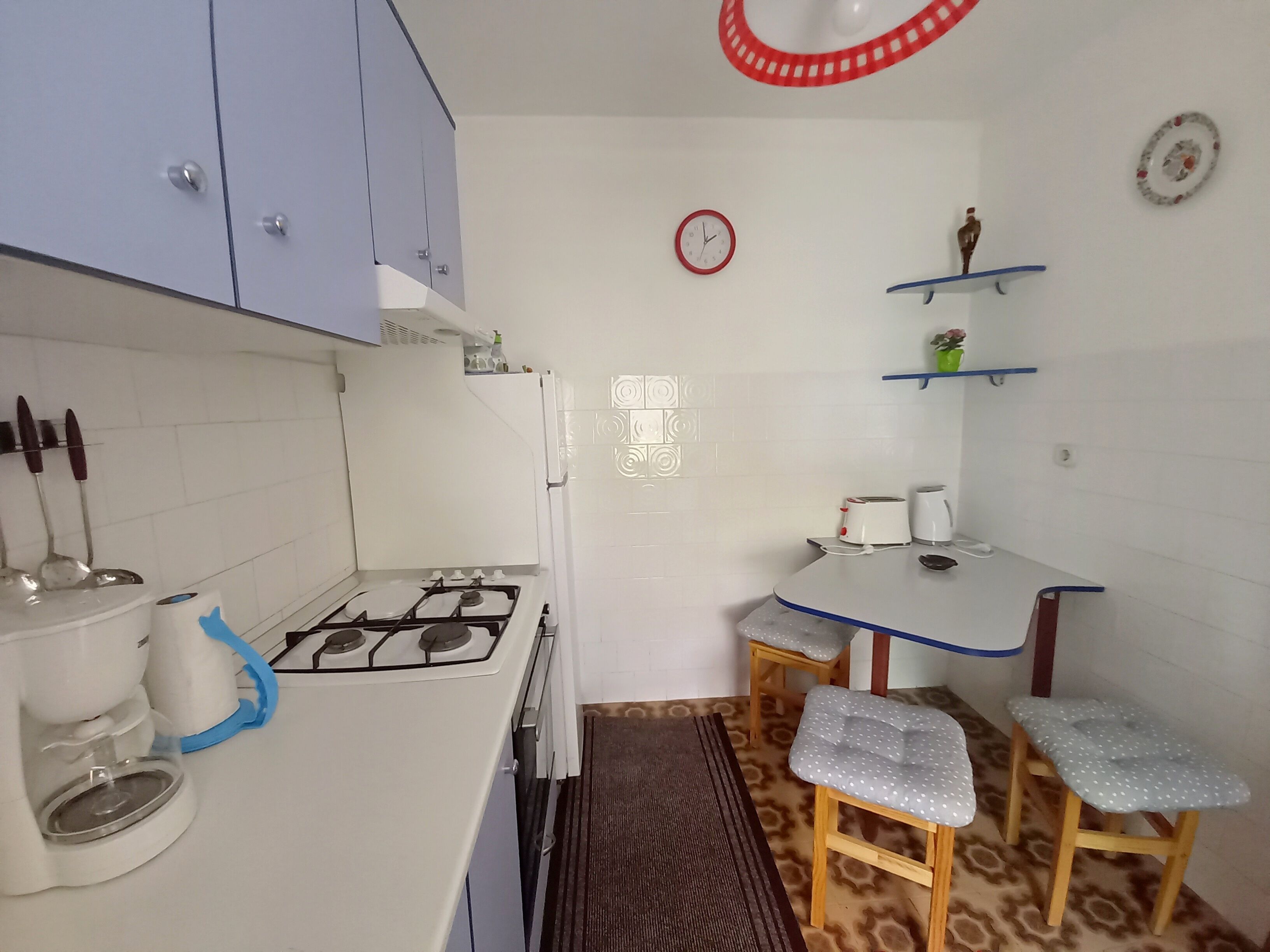 Cocina compacta con estufa a gas, cafetera y lavamanos. Pequeño comedor con dos taburetes y una mesa.