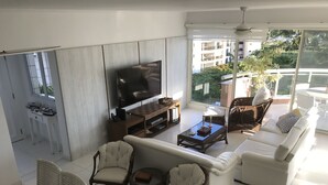 A modern living room with a flat-screen TV, white sofas, wicker chairs, and a coffee table.