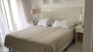 A large bed with a beige bedspread, two bedside tables with lamps, and a view of the outdoors through a window with sheer curtains.