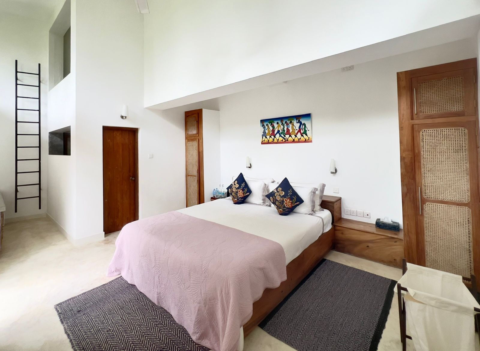 A bedroom with a bed, pillows, a nightstand, a lamp, a painting, a wooden door, a window, and a ladder.