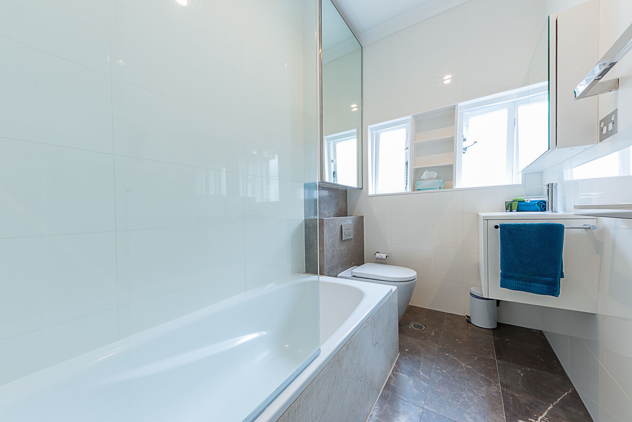 Une salle de bain moderne avec une baignoire entourée de parois en verre, des toilettes et un lavabo agrémenté d’une serviette bleue.