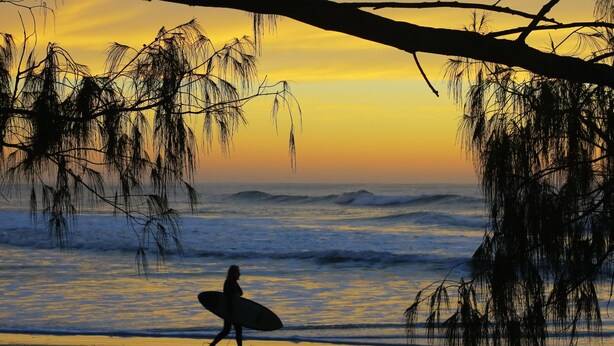 Escena de playa con una persona cargando una tabla de surf, olas al fondo y un cielo al atardecer.