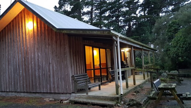 A wooden cabin with a porch, benches, and a covered outdoor seating area. - Rustic cabin & large kitchen block nestled in a native garden. (Western Lake, Featherson)