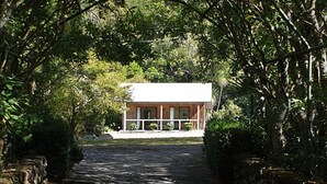 A house with a white exterior and a porch, surrounded by dense greenery and trees. - Rustic cabin & large kitchen block nestled in a native garden. (Western Lake, Featherson)