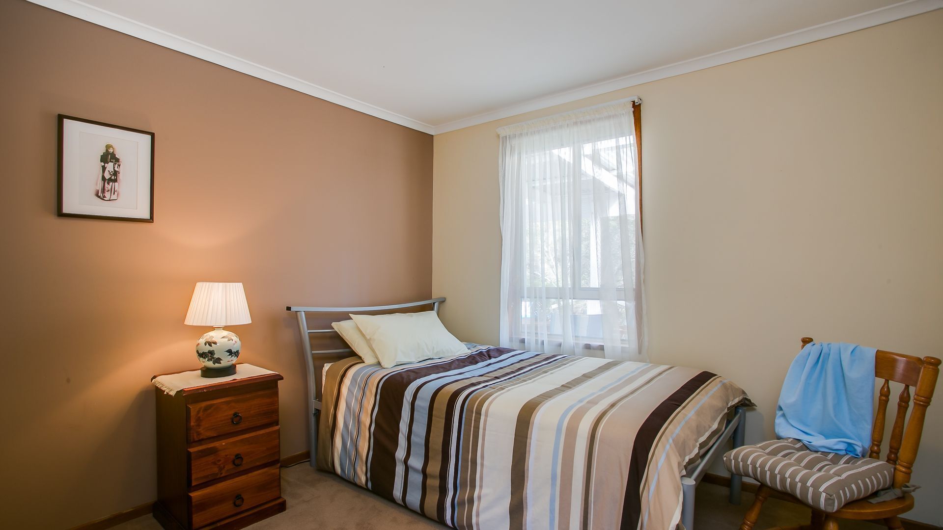 A bedroom with a bed, a nightstand with a lamp, a chair, and a window with sheer curtains.