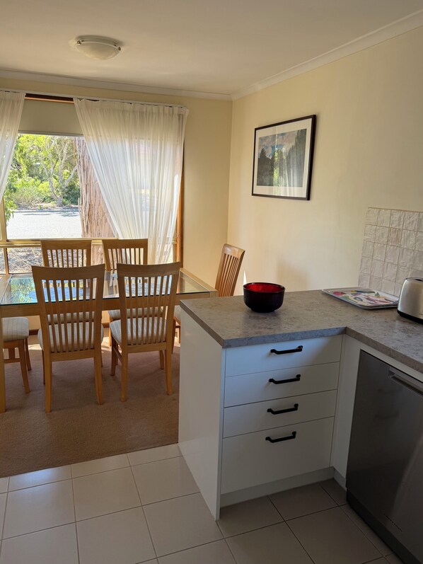 Een keuken met een eethoek, met houten stoelen, een glazen tafel, een wit kastje met lades, een zwarte kom en een ingelijste foto aan de muur.