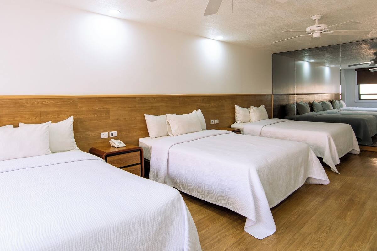 A hotel room with three beds, wooden headboards, a wooden floor, and a ceiling fan.