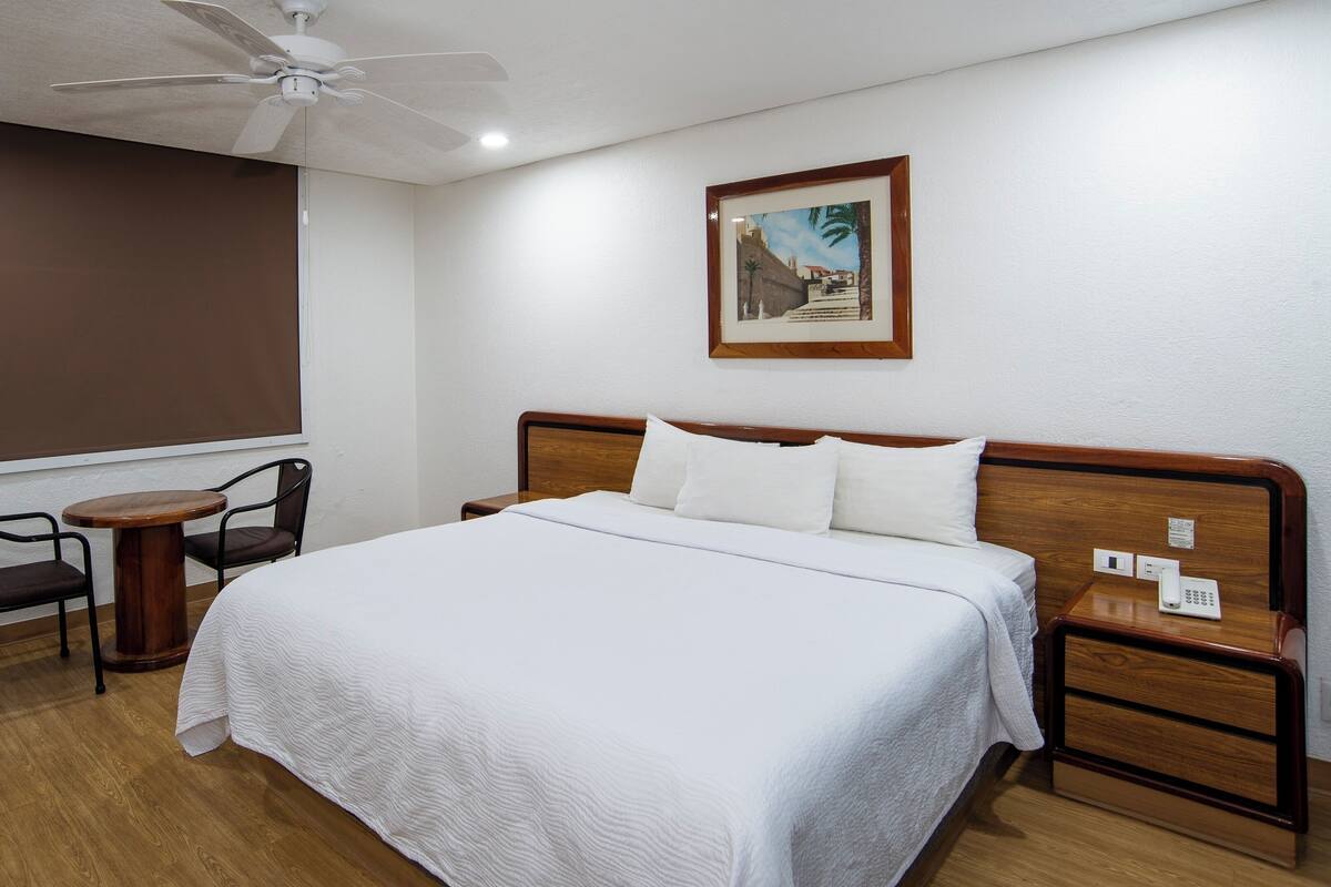 A hotel room with a bed, a bedside table, a chair, a ceiling fan, and a window with a view of buildings.
