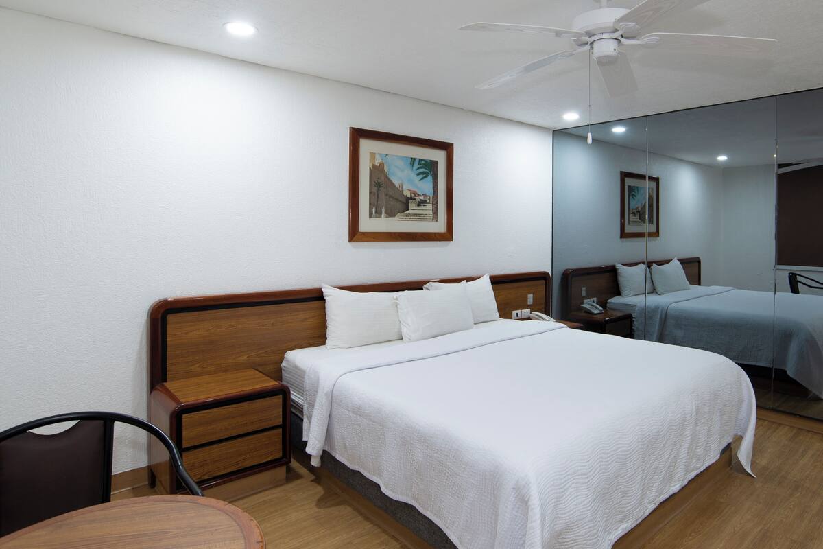 A hotel room with two beds, wooden headboards, a small table, a chair, a nightstand, a phone, and a ceiling fan.