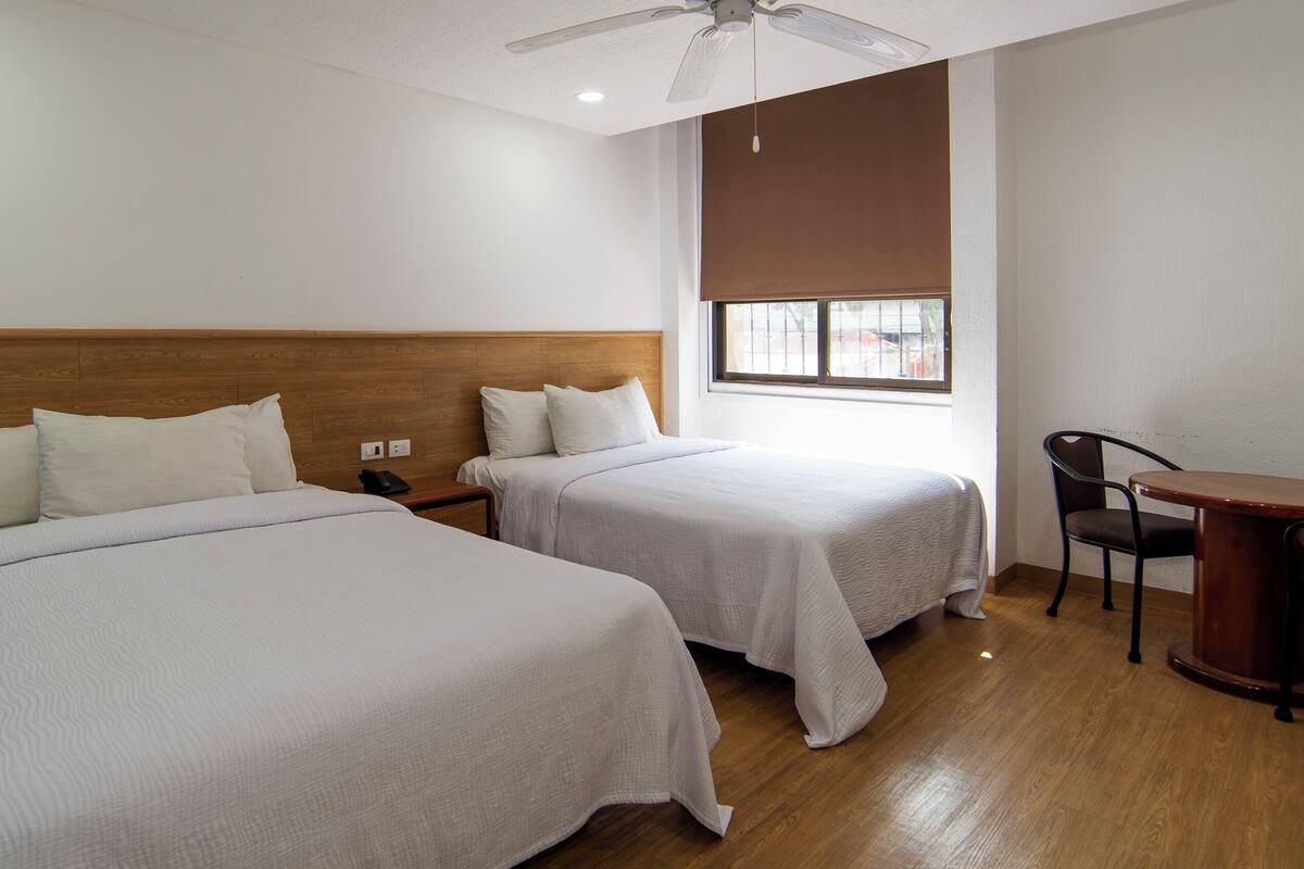 A hotel room with two beds, a wooden headboard, a desk with a chair, a window with blinds, and a ceiling fan.