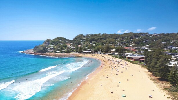 A beach with clear blue waters, sandy shore, and houses on a hillside.