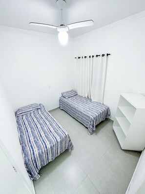 A room with two beds, a ceiling fan, white walls, and a white shelving unit.