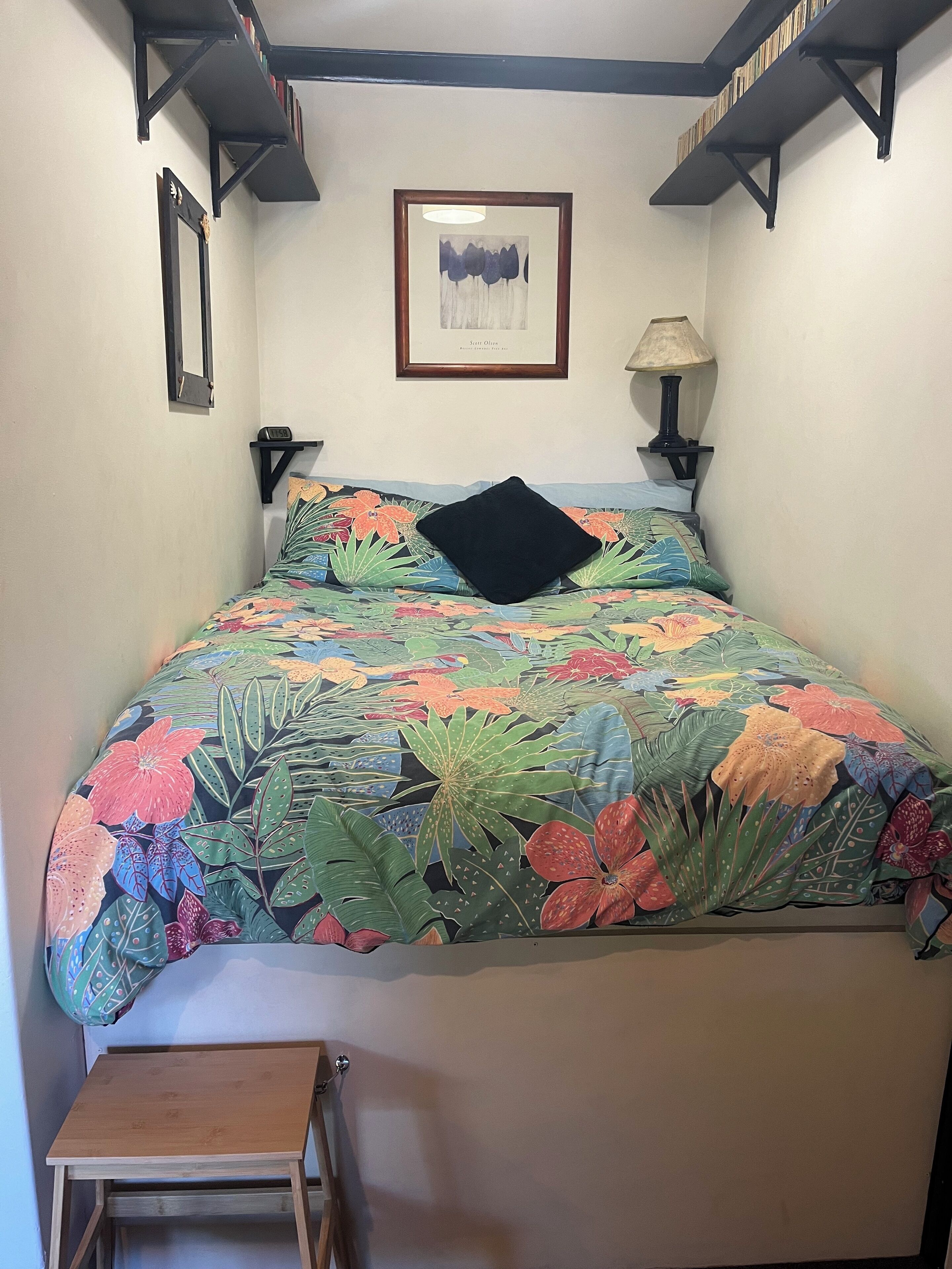 A bed with a floral bedspread, a black pillow, a framed picture on the wall, and a small wooden table beside the bed.