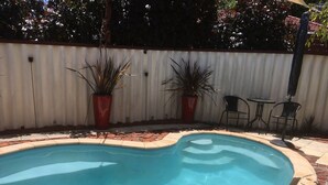 A swimming pool with a curved edge, surrounded by a brick border and a white fence. There are two potted plants and a small table with chairs.