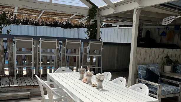 A covered outdoor dining area with a white table and chairs, a grill, and a view of the sky through a glass roof.