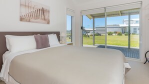 Un grand lit avec une literie blanche et marron, une table de chevet avec une lampe, et une vue sur une maison moderne et une voiture, visibles à travers une porte-fenêtre coulissante.