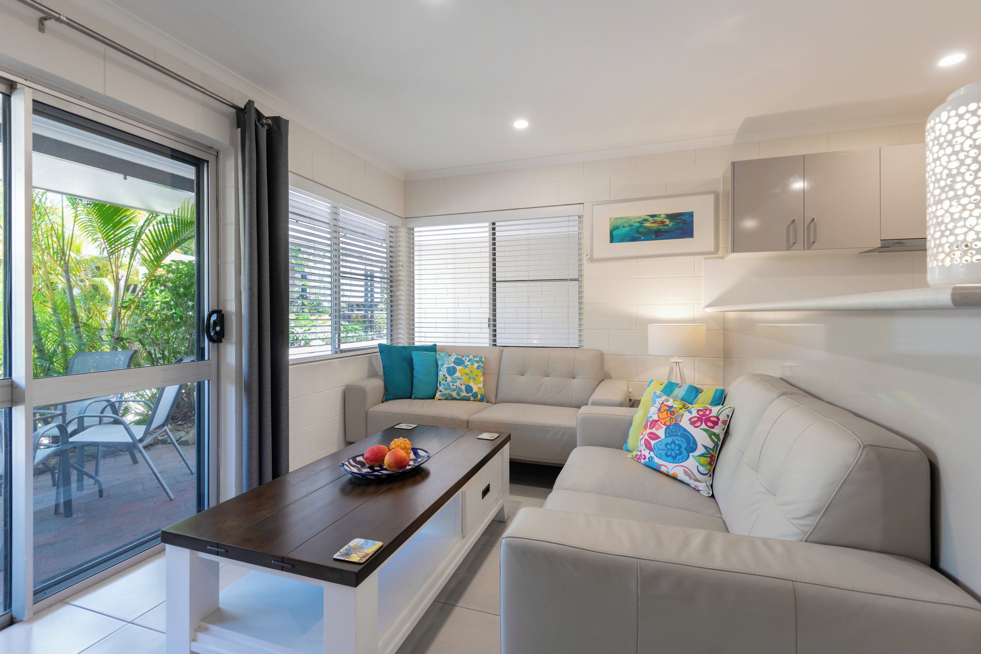 A modern living room with a sofa, a coffee table with fruit, and a view of greenery through large windows.