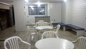 Une petite pièce carrelée, aux murs blancs et au sol carrelé. On y trouve plusieurs chaises en plastique blanc et une petite table ronde au centre. Une fenêtre est visible sur le mur du fond.