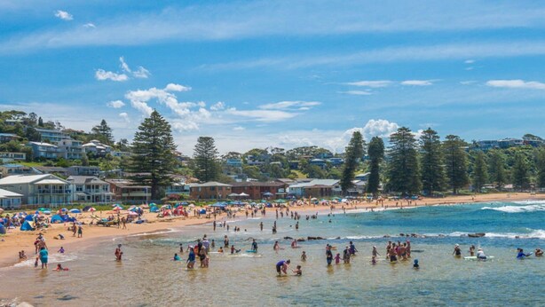 A beach with people swimming and sunbathing, houses in the background, and clear skies.