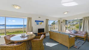 A room with a large window overlooking the beach, wicker furniture, a television, and a painting.