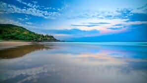 A beach with a calm sea, clear sky, and a forested hillside.