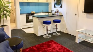 A modern kitchen with white cabinets, a blue backsplash, and a red pendant light. There are two blue bar stools and a small island with a sink. - Amber Homes B Half Moon Bay (Bucklands Beach)