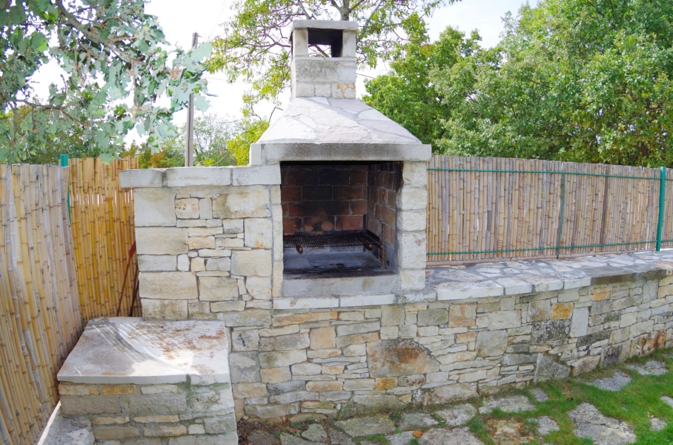 A stone-built outdoor oven with a chimney, surrounded by a bamboo fence and a stone wall.