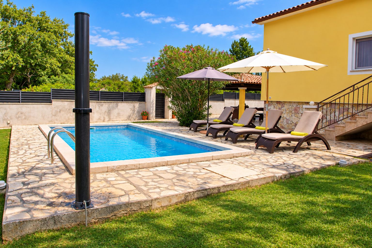 Una piscina con sillones de descanso y sombrillas, rodeada por un patio de piedra y un edificio amarillo con una escalera.
