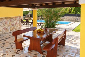Eine Terrasse im Freien mit Steinplattenboden, Holzbanken, einem Tisch mit Früchten und einer Flasche, und einem Pool im Hintergrund.