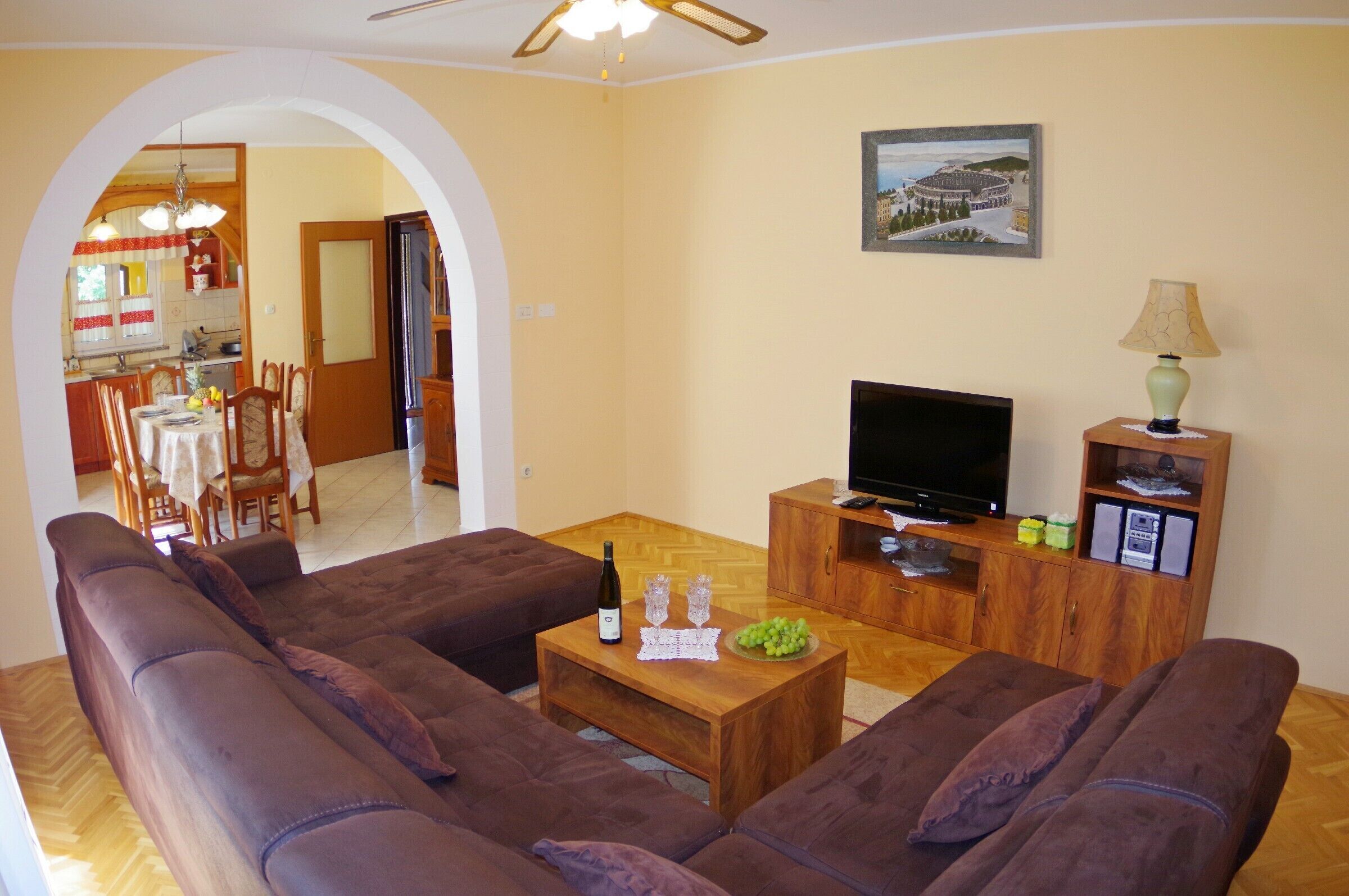 A living room with a sofa set, a coffee table with a bottle and glasses, a television on a wooden cabinet, and a dining area visible through an archway.