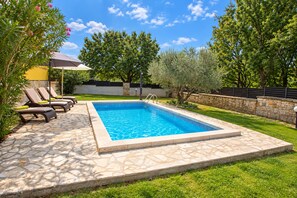 Una piscina rectangular con borde de piedra, rodeada de césped y árboles.