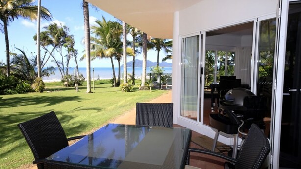 A patio with a glass table and chairs, overlooking a lawn with palm trees and a view of the ocean.
