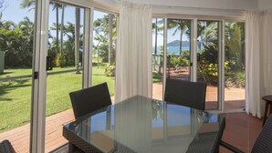 A glass-top table with wicker chairs in a room with large sliding glass doors leading to a patio with a view of palm trees and a body of water.