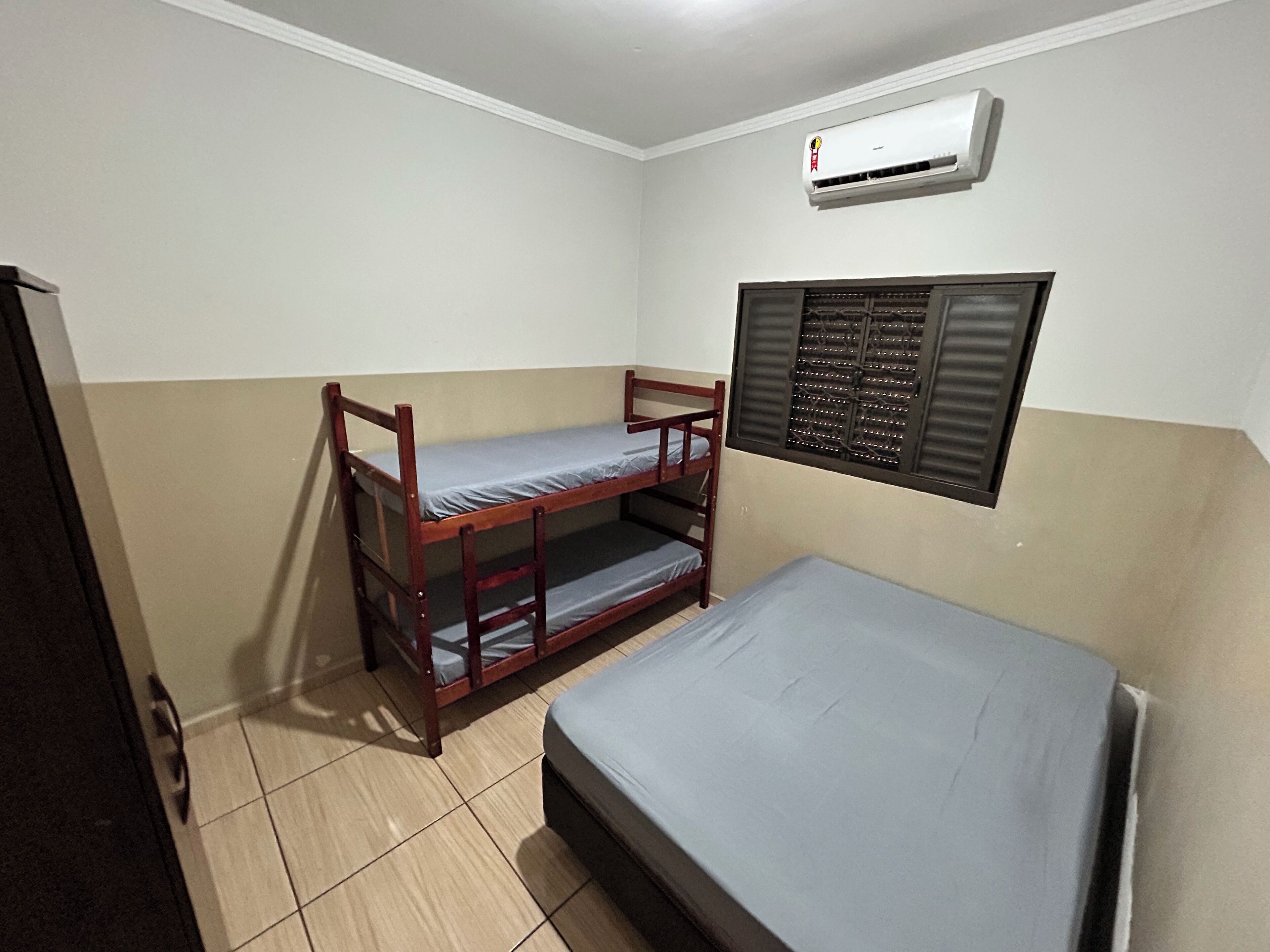 Een kamer met een stapelbed, een enkel bed, een airconditioning en een raam met jaloezieën.