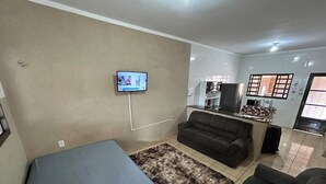 Ein Wohnzimmer mit einer Couch, einem an der Wand montierten Fernseher, einer Küchenzeile mit Kühlschrank und Kaffeemaschine sowie einem kleinen Teppich.
