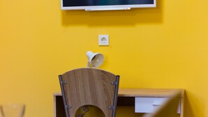 Een kamer met een gele wand, een wandgemonteerde flatscreen-tv, een houten bureau, een stoel en een ronde tafel.
