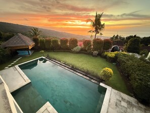 Une piscine avec vue sur le coucher du soleil, une construction au toit de chaume et des haies bien entretenues.