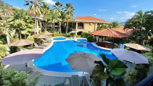 A swimming pool with multiple sections, surrounded by palm trees and umbrellas, with a building in the background. - Great second floor condo with pool view- Best location in Jaco beach (Jaco)