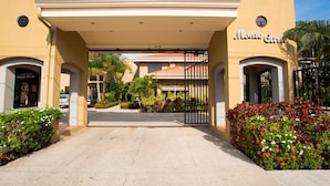 L’entrée d’un hôtel, avec un passage couvert, des palmiers et une allée bordée de grilles.