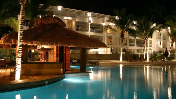 A pool area with a thatched-roof structure, palm trees, and a multi-story building with balconies. - Ground floor, Poolside Luxury Apartment (Palm Cove)