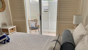 A bedroom with a bed, pillows, a chair, a nightstand, and a window with blinds. - Ground floor, Poolside Luxury Apartment (Palm Cove)
