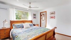 Une chambre à coucher avec un lit en bois, une table de chevet, une lampe de bureau, un ventilateur de plafond et une fenêtre avec des rideaux.